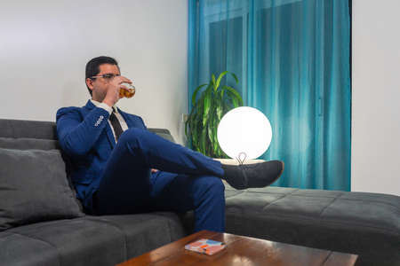 Young man sitting on a sofa with his legs crossed. He is wearing a blue suit. He drinks a glass of whiskey.の写真素材