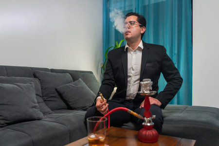 Young man sitting on a sofa. I have smoking with a shisha. The hookah is red. On a wooden table...