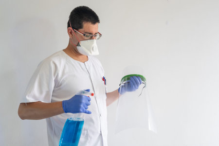 Doctor / nurse cleaning a face shield. He wears gloves and a mask. His uniform is white.