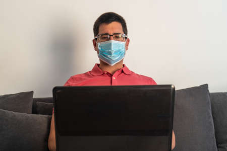 Man with a mask working on his laptop from home. He is sitting in an armchair. He is wearing a salmon colored polo shirt and glasses.の写真素材