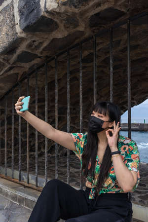 Young Asian tourist woman taking a selfie with her smartphone. She has a floral shirt and mask. Puerto de La Cruz is a tourist site in Tenerife.の写真素材