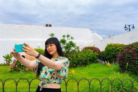 Young Asian tourist woman taking a selfie with a Canarian garden behind. He wears a floral shirt and...