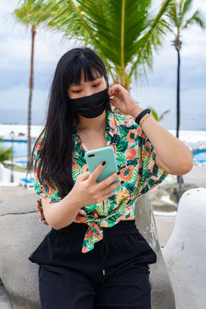 Young Asian tourist chatting with her smartphone. It is located in the pool of Lago Martianez, in Puerto De La Cruz in Tenerife. She wears a floral shirt and mask.の写真素材