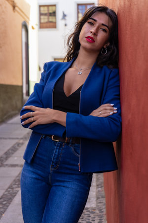 Young woman with arms crossed leaning against a red wall. She wears jeans and an American jacket. The photo was taken in La Laguna, Tenerife.の写真素材