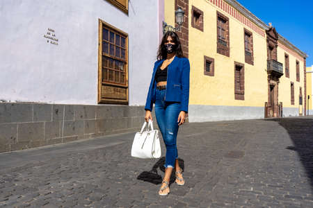 Young woman walking down a pedestrian street in La Laguna, Tenerife. She wears mask, jeans, an American jacket, a bag and sandalsの写真素材