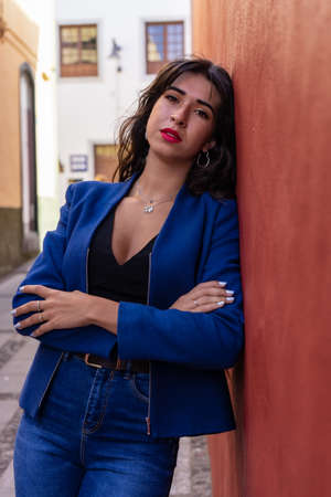 Young woman with arms crossed leaning against a red wall. She wears jeans and an American jacket. The photo was taken in La Laguna, Tenerife.の写真素材