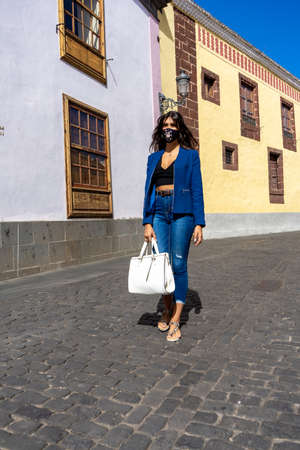 Young woman walking down a pedestrian street in La Laguna, Tenerife. She wears mask, jeans, an American jacket, a bag and sandalsの写真素材