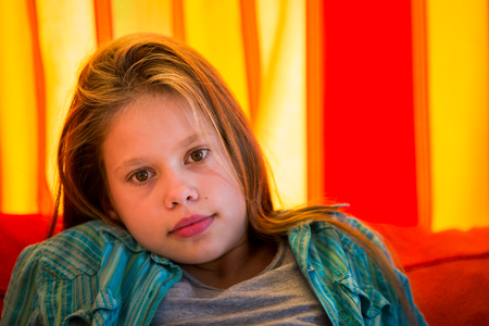 Head shot of a beautiful, 11 years old girl sitting in front of a red and orange curtainの写真素材