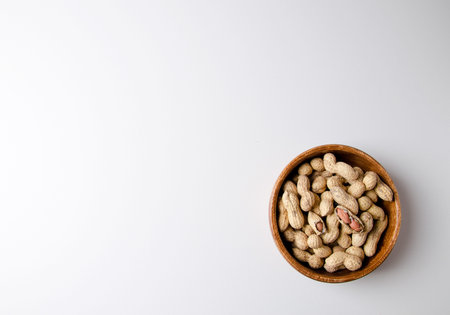 Unpeeled peanuts lies in a wooden bowl on a white background. On top, one nut is half peeled. Flat...
