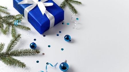 Gift box in blue paper with white satin ribbon on light background with blue christmas balls with...