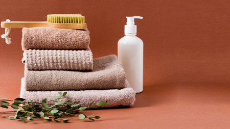 A stack of clean cotton brown towels with a body massage brush and a bottle of cosmetic body cream...