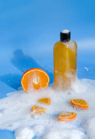 Shampoo, gel, liquid soap of orange color from natural ingredients in a transparent bottle stands on a blue background in soapy foam with orange pieces, vertical background, shadows, copy spaceの写真素材