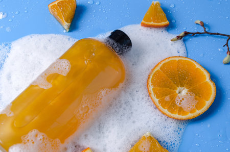 Shampoo, gel, liquid soap of orange color from natural ingredients in a transparent bottle on a blue background in soapy foam with orange pieces, flat lay copy spaceの写真素材