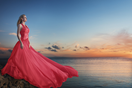 Beautiful young woman posing in red luxurious long dress on the sea beachの写真素材