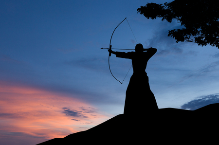 Abstract photo of man silhouette demonstrating martial arts with bow in front of sunset skyの写真素材