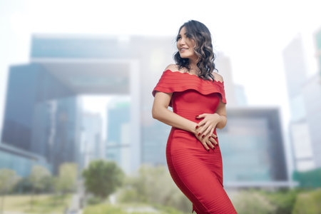 An elegant Asian woman in a stunning red dress striking a pose in front of modern office buildings in a bustling metropolis. Ideal for fashion, urban lifestyle, and cityscape concepts.の写真素材