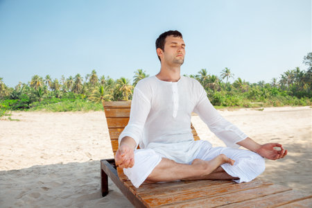 A young adult man peacefully relaxing and meditating on a sunny tropical beach, surrounded by serene beauty. Ideal for wellness, travel, and relaxation concepts.の写真素材