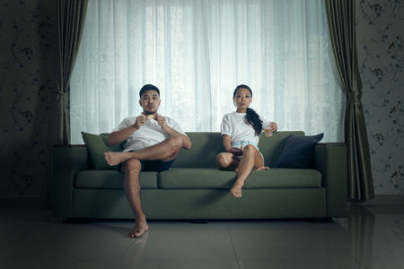 A young couple dressed casually sits on a green couch in a living room against a curtained background, gazing pensively.の写真素材