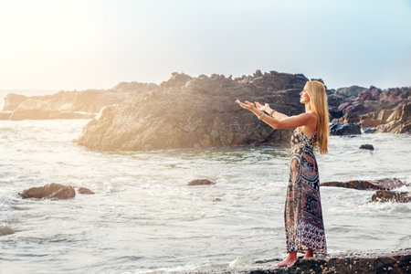 Woman in a Dress by the Rocky Shore. Young woman with long blonde hair on a rocky shore, gazing at the horizon. A tranquil and picturesque scene.の写真素材