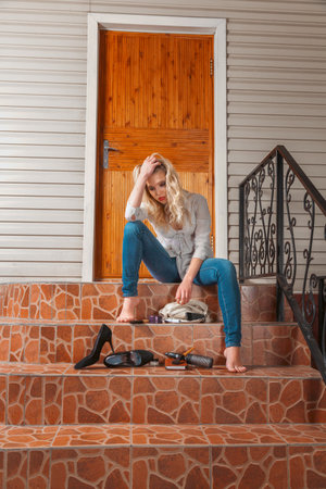 Sad young woman sits in front of closed entrance door because she lost her keys, appearing frustrated and disheveled. A moment of distress and inconvenience. Locksmith services business.の写真素材