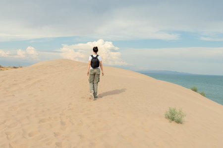 A lone hiker with backpack stands atop a sand dune, facing a vast ocean view. Perfect for travel blogs, adventure tourism ads, or articles on solo exploration and natural landscapes.の写真素材