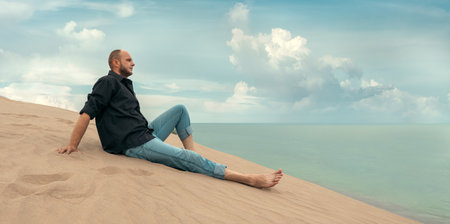 A man sits peacefully on a sand dune, gazing at the calm ocean and sky. Perfect for wellness articles, meditation apps, or travel content focusing on relaxation and self-reflection.の写真素材