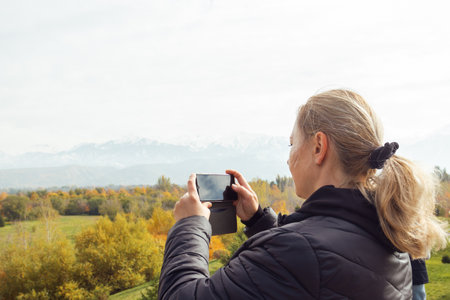 Blonde woman capturing scenic fall view of mountains and colorful foliage with her phone. for travel, nature photography, or outdoor lifestyle content.の写真素材