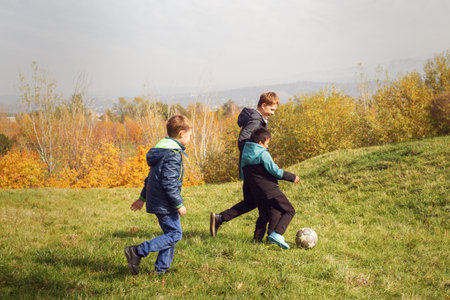 Young boys kicking football on grassy hill surrounded by fall foliage. Perfect for themes of outdoor play, childhood friendships, seasonal activities, or active lifestyle concepts.の写真素材