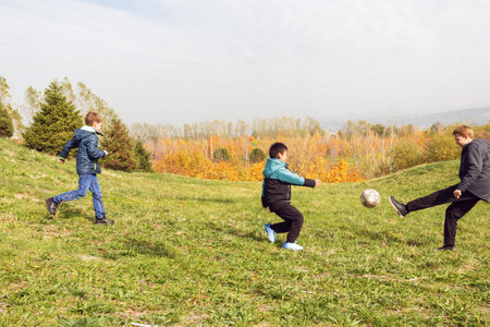 Young boys kicking football on grassy hill surrounded by fall foliage. Perfect for themes of outdoor play, childhood friendships, seasonal activities, or active lifestyle concepts.の写真素材