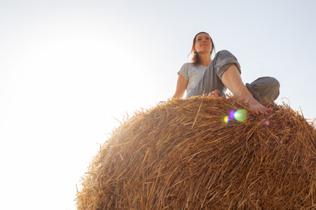 Woman in peaceful meditation pose atop hay bale in vast rural field. Countryside tranquility, mindfulness practice, and connection with nature. Wellness, stress relief, or rural lifestyle concepts.の写真素材