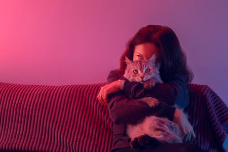 a brunette woman embracing her cat on a red couch, bathed in pink and purple neon light. for themes of pet companionship, relaxation at home, or modern lifestyle imagery.の写真素材