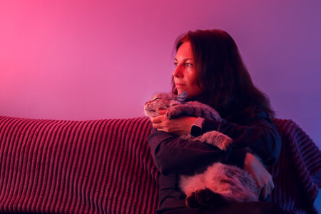 a brunette woman embracing her cat on a red couch, bathed in pink and purple neon light. for themes of pet companionship, relaxation at home, or modern lifestyle imagery.の写真素材