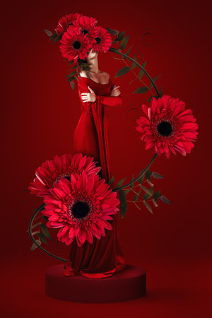 Artistic fashion photograph featuring model in flowing red dress with oversized gerbera daisy blooms on monochromatic red backdrop. for fashion editorials, beauty campaigns or luxury advertising.の写真素材
