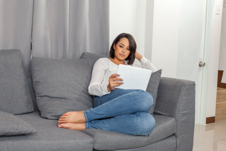 Woman using tablet device while relaxing on comfortable sofa. for lifestyle, technology, remote work, digital entertainment, and home comfort concepts.の写真素材