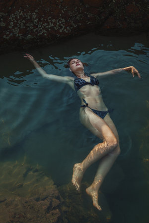 Young woman in blue bikini floating peacefully in dark water of natural swimming hole. for travel, vacation, wellness, relaxation, and summer lifestyle concepts.の写真素材