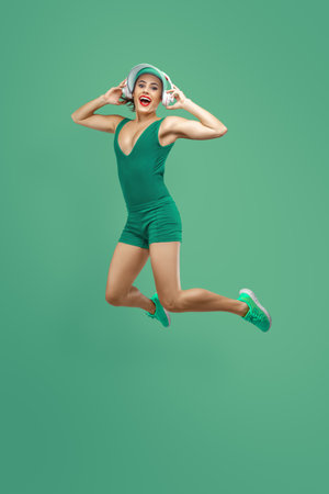 A joyful young woman in green sportswear and headphones jumps mid-air. for advertising, lifestyle, fitness, music, and emotional well-being concepts.の写真素材