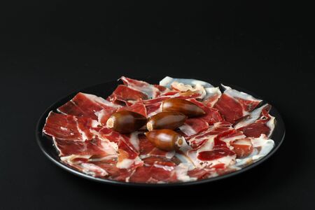 Ration of acorn-fed Iberian ham on a black plate with acorns garnishing. The background is black and the image has selective focusの写真素材