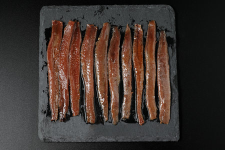 Portion of anchovies from SantoÃ±a on a square slate plate viewed from above and black background