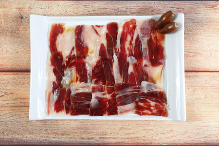 Portion of acorn-fed Iberian ham in a white rectangular tray, on a rustic wooden table and decorated...