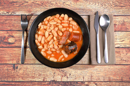 Asturian fabada (white beans) on black plate with napkin and cutlery on rustic wooden tableの写真素材
