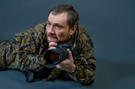 Bearded camera man dressed in camouflage uniform lying on the floor hunts his target with camera in...