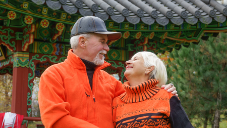 Happy beautiful elderly married couple with gray hair in orange sportswear on a background of autumn trees with yellow leavesの写真素材