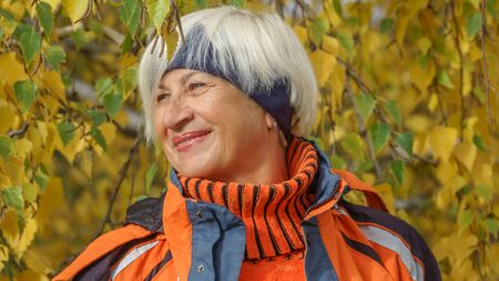 Happy beautiful elderly woman with gray hair in sportswear on a background of autumn trees with yellow leavesの写真素材