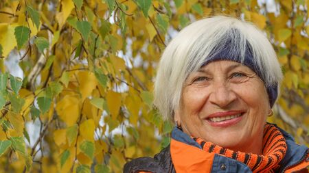 Happy cute elderly woman with gray hair in sportswear on a background of autumn trees with yellow leavesの写真素材