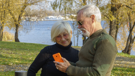 Portrait of a loving elderly couple with a smartphone in hand on the background of the autumn city parkの写真素材