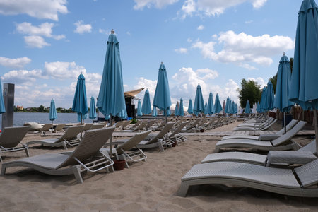 Blue closed stationary beach umbrellas and sun loungers on a sandy beach on the bank of a river...
