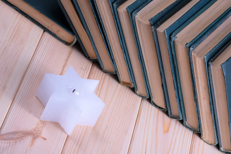 A white paraffin candle shaped like a six-pointed Jewish star stands next to a stack of green-bound books on a wooden tableの写真素材