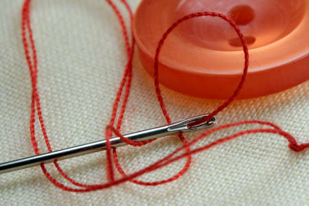 Red button with a needle with inserted thread on a white linen cloth close-up macro photographyの写真素材