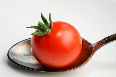 Small red ripe tomato in a teaspoon on a white mirror background close-up