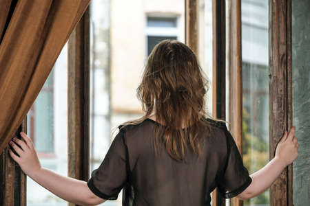 A young pretty attractive brown-haired girl in a transparent black peignoir stands with her back next to the open door to the balcony close-upの写真素材